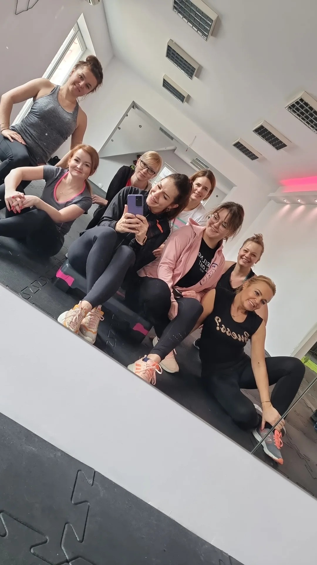 Grupa uśmiechniętych kobiet w strojach sportowych pozuje do selfie w lustrze na tle sali gimnastycznej, widać stepy i maty podłogowe.