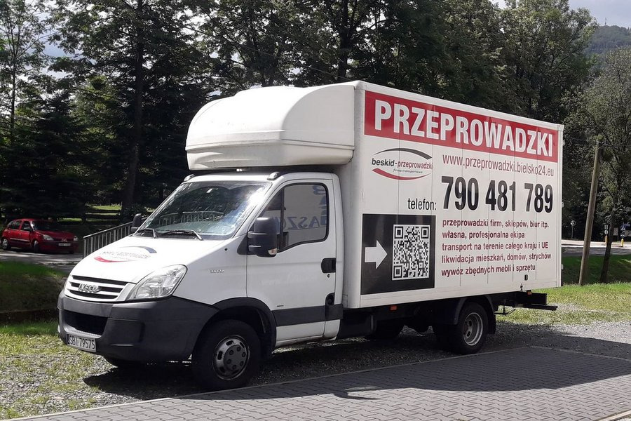 Biała furgonetka z czerwonym napisem 'PRZEPROWADZKI' i numerem telefonu 790 481 789, zaparkowana na parkingu z drzewami w tle.