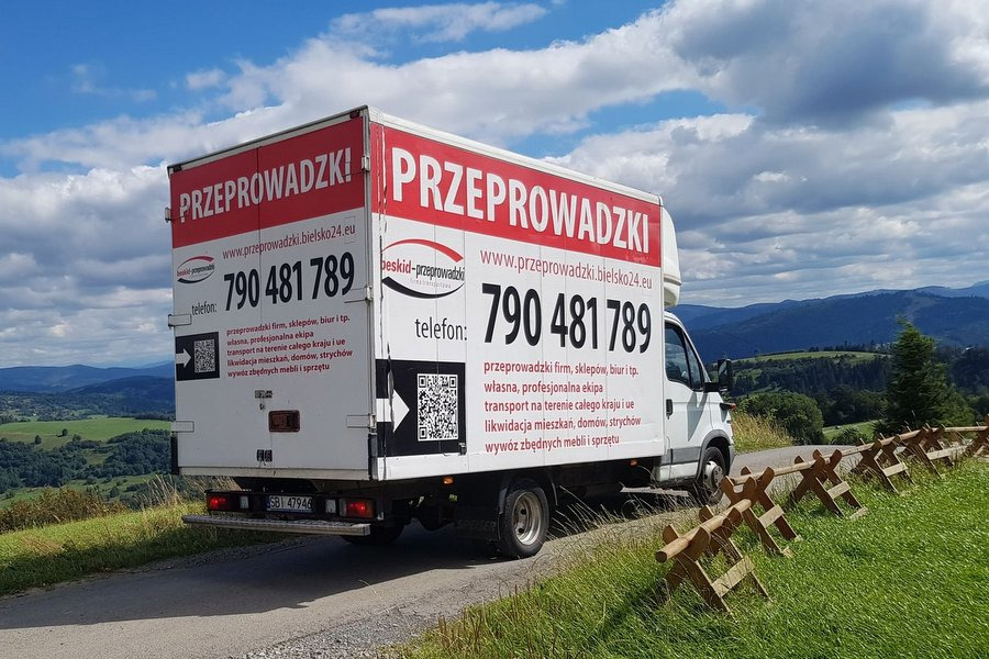 Samochód dostawczy z reklamą firmy przeprowadzkowej na tle górskiego krajobrazu, zaparkowany obok drewnianego płotku.