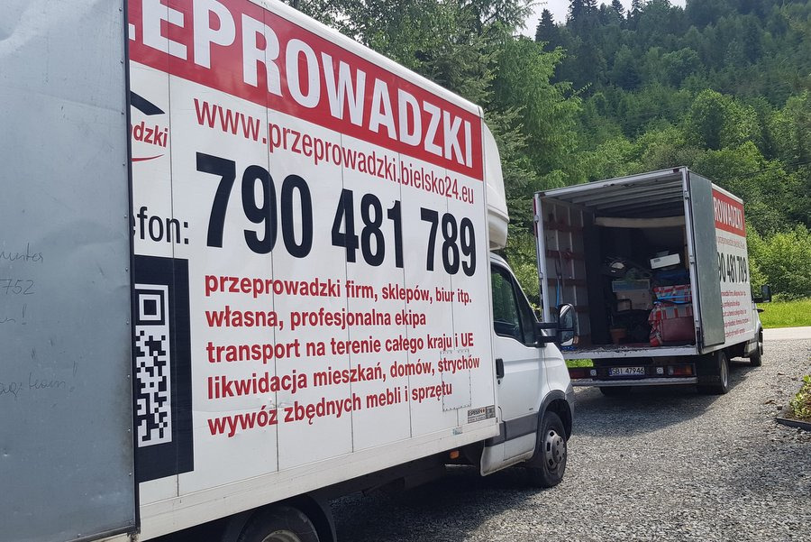 Dwa samochody dostawcze z napisem 'Przeprowadzki' zaparkowane na żwirowej drodze, z otwartym tyłem jednego z nich, ukazującym zapakowane przedmioty, na tle zielonego lasu.