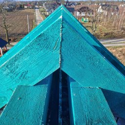 Usługi blacharsko-dekarskie JAN-DEK - Widok z góry na fragment konstrukcji dachu pokrytej turkusową impregnacją, widoczne deski tworzące szkielet dachu na tle wiejskiej zabudowy i pola.