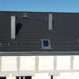 Nowy, ciemnoszary dach z dachówki ceramicznej z trzema oknami dachowymi i dwoma kominami, widoczny na tle częściowo wykończonej ściany budynku z białych bloczków.