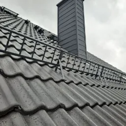 Szary dach z ceramicznej dachówki karpiówki z zamontowaną metalową barierką śniegową i obróbką komina z blachy na tle pochmurnego nieba.