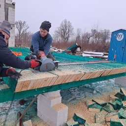 Usługi blacharsko-dekarskie JAN-DEK - Dwóch pracowników tnie deskę na zielonej konstrukcji za pomocą piły tarczowej, w tle niezadaszony budynek w trakcie budowy i przenośna toaleta.