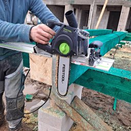 Usługi blacharsko-dekarskie JAN-DEK - Pracownik w ubraniu roboczym tnie belkę drewnianą piłą łańcuchową Festool Univers SSU 200 EB na budowie, widoczne zielone impregnowane elementy konstrukcji dachu i błoto.