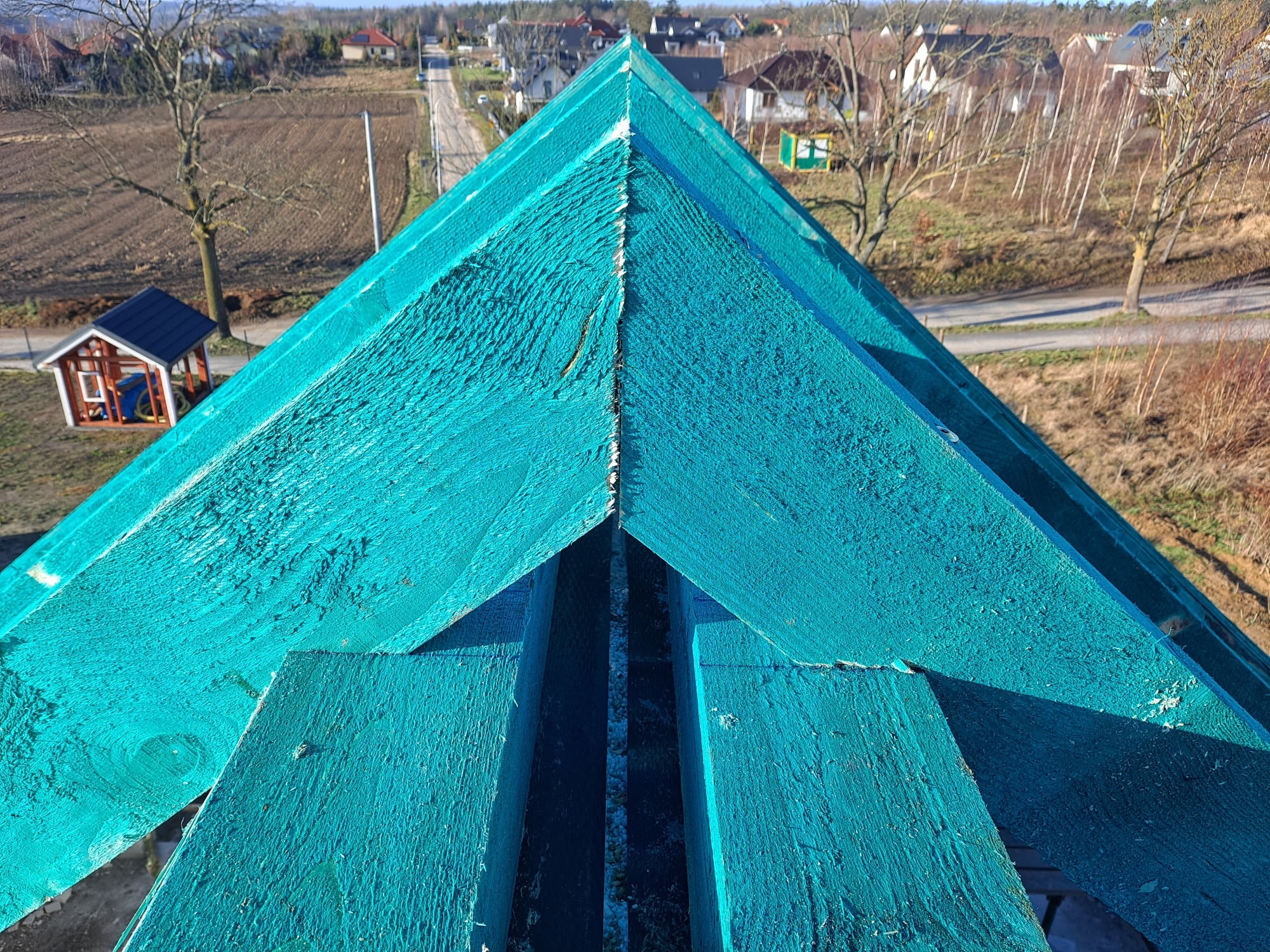 Widok z góry na fragment konstrukcji dachu pokrytej turkusową impregnacją, widoczne deski tworzące szkielet dachu na tle wiejskiej zabudowy i pola.