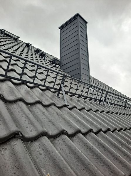 Szary dach z ceramicznej dachówki karpiówki z zamontowaną metalową barierką śniegową i obróbką komina z blachy na tle pochmurnego nieba.