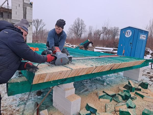 Dwóch pracowników tnie deskę na zielonej konstrukcji za pomocą piły tarczowej, w tle niezadaszony budynek w trakcie budowy i przenośna toaleta.