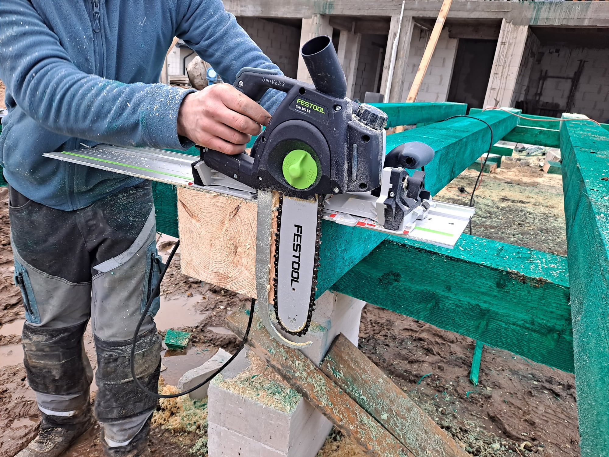 Pracownik w ubraniu roboczym tnie belkę drewnianą piłą łańcuchową Festool Univers SSU 200 EB na budowie, widoczne zielone impregnowane elementy konstrukcji dachu i błoto.