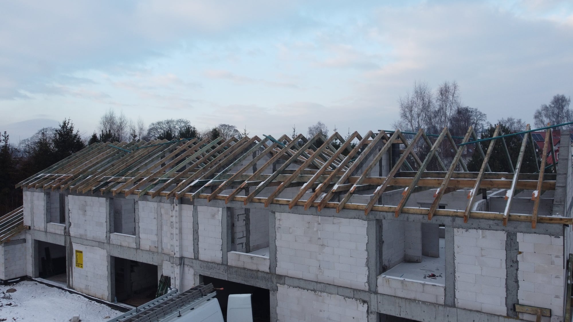 Konstrukcja drewniana dachu w trakcie budowy, widok z góry na surową więźbę na domu z białych bloczków, zimowy krajobraz w tle.