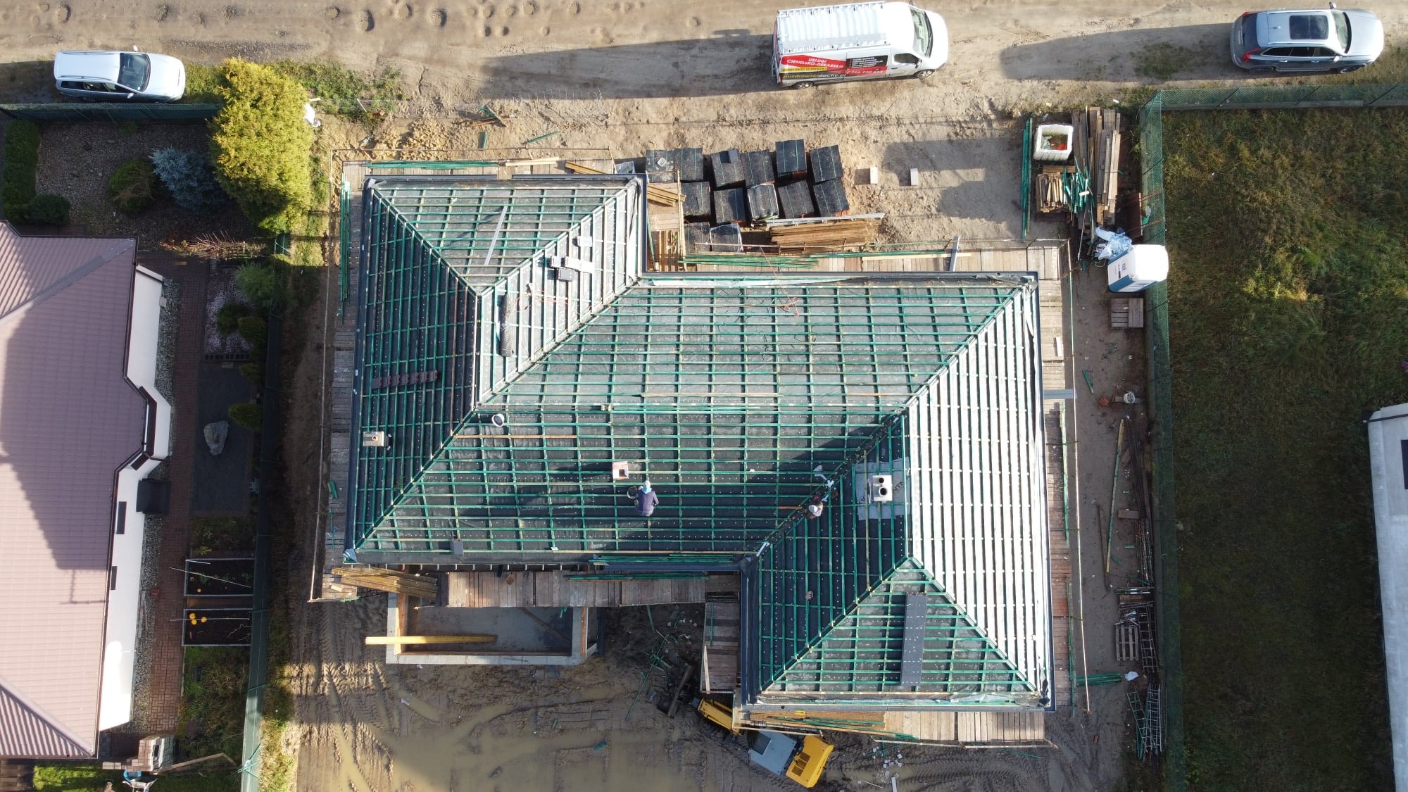 Widok z góry na budowę dachu domu jednorodzinnego, z widoczną konstrukcją więźby dachowej, czarną membraną i pracującymi dekarzami, na ziemi leżą materiały budowlane i stoi żółta koparka.