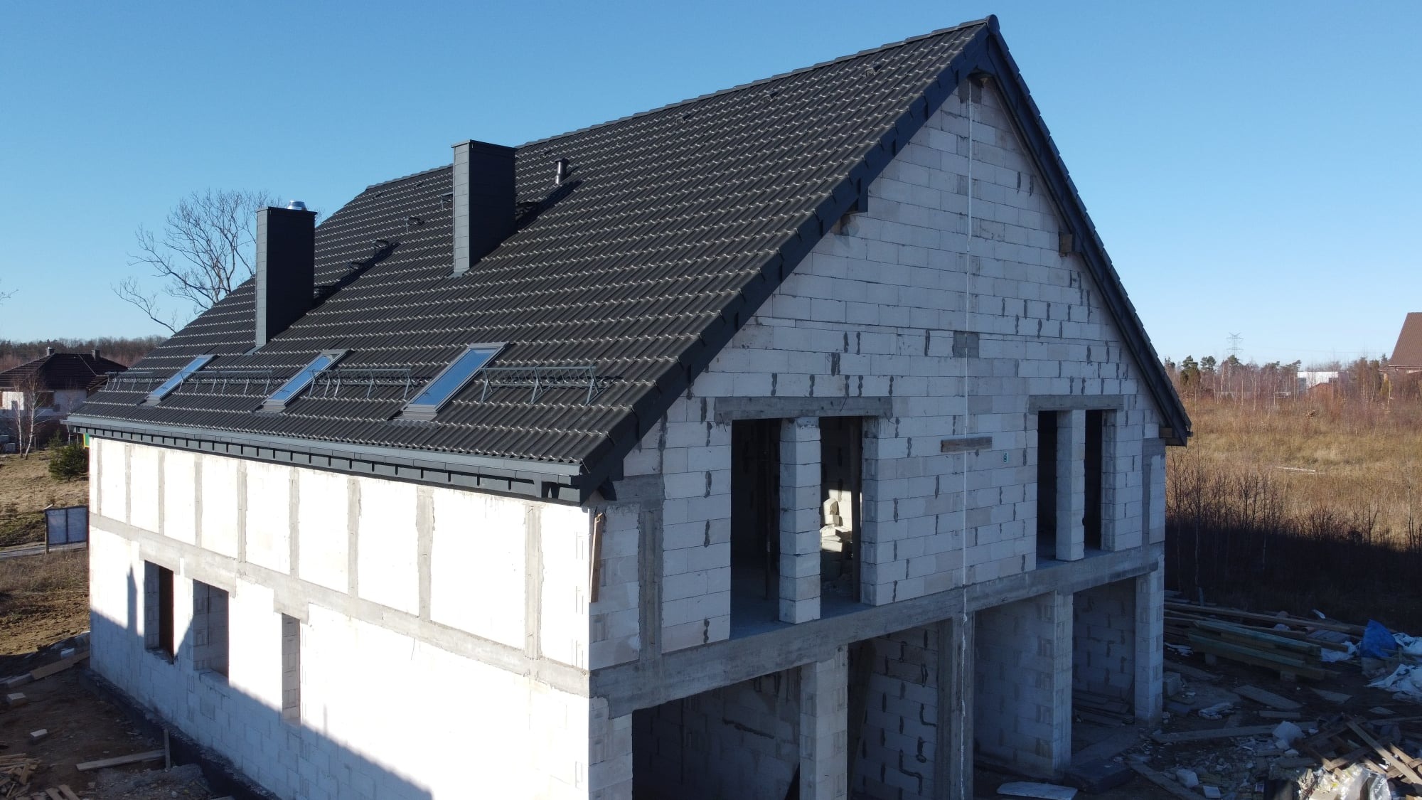 Niedokończony dom dwukondygnacyjny z szarym dachem pokrytym dachówką ceramiczną, widoczne okna dachowe i kominy, ściany z białych bloczków.