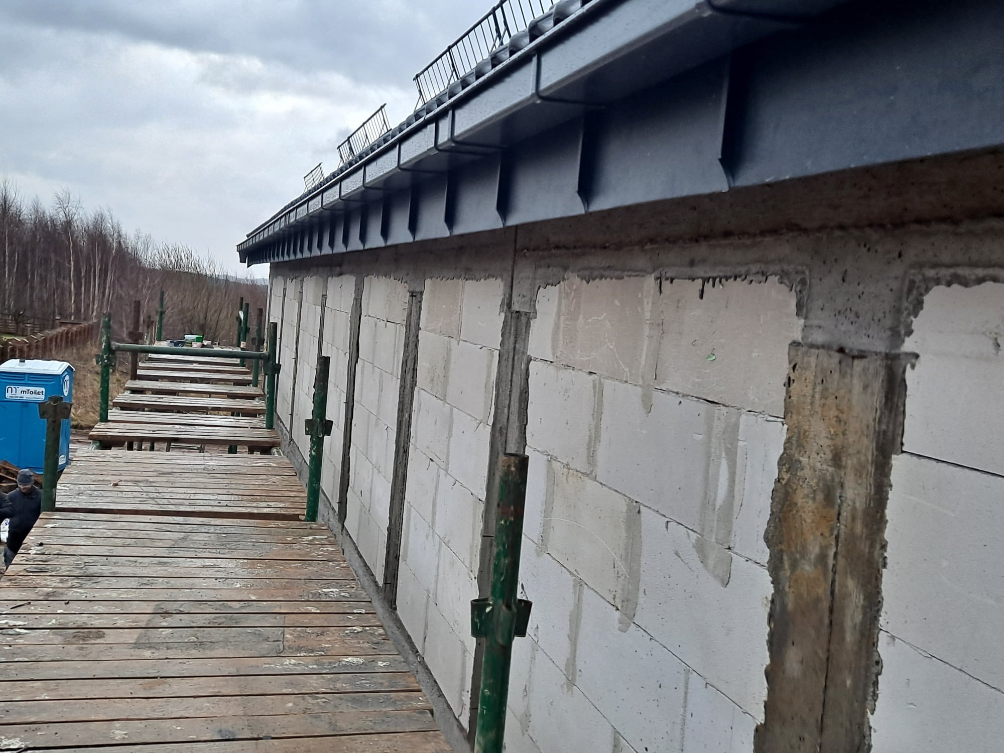 Surowy stan budowy: Widok z rusztowania na fragment ściany budynku z betonu komórkowego, z zamontowaną czarną, metalową rynną i elementami dachu, na tle pochmurnego nieba i drzew bez liści.