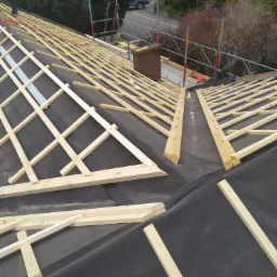 Konstrukcja drewniana dachu pokrytego czarną membraną, przygotowana pod krycie dachówką, widok z góry na tle zieleni i zabudowań.