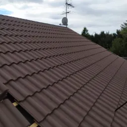 Fragment brązowego dachu z dachówki ceramicznej, widoczne drewniane elementy konstrukcji, w tle antena telewizyjna i pochmurne niebo.