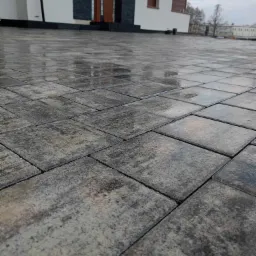 Szare, prostokątne płyty brukowe ułożone przed nowoczesnym domem z ciemnymi oknami i drzwiami wejściowymi, lekko mokre od deszczu.