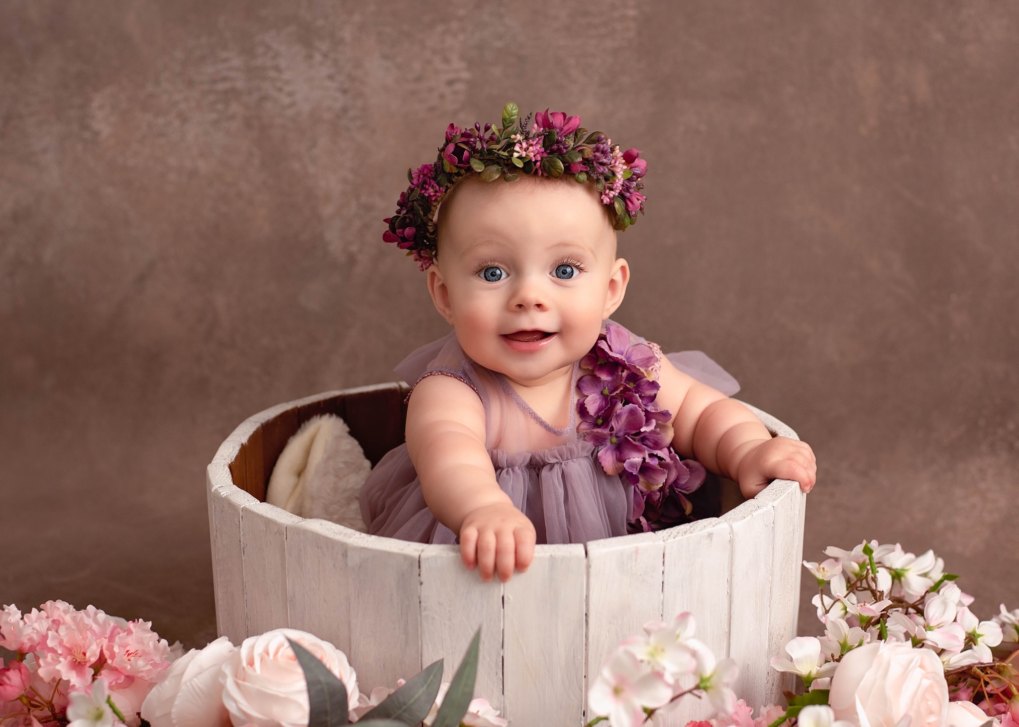 Uśmiechnięta niemowlęca dziewczynka z niebieskimi oczami w fioletowej sukience i wianku z kwiatów na głowie, siedząca w białym, drewnianym wiadrze otoczonym różowymi i białymi kwiatami.