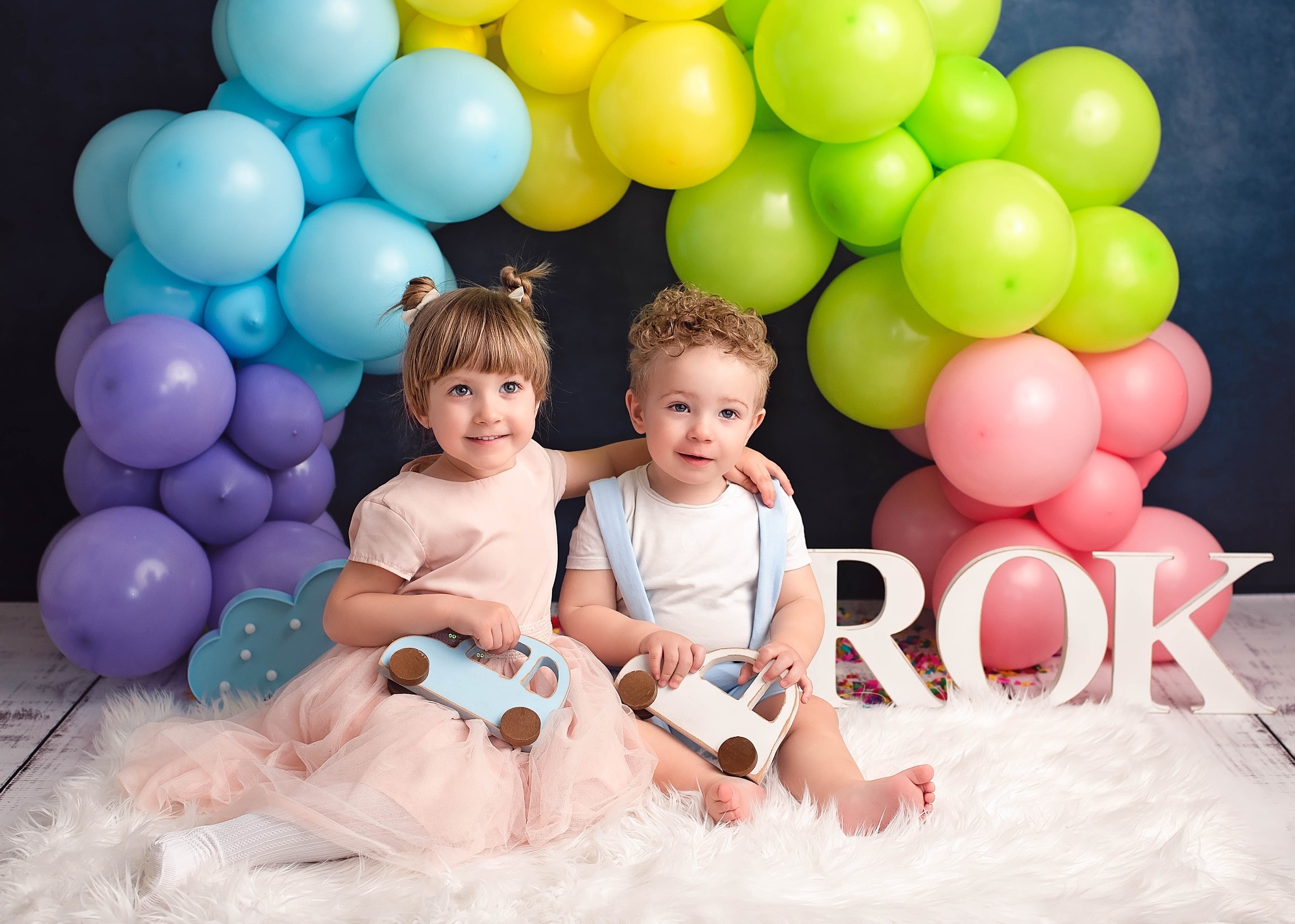 Uśmiechnięta dziewczynka w tiulowej sukience i chłopiec z kręconymi włosami siedzą na puszystym dywanie w Rybniku, trzymając drewniane zabawki w kształcie samochodów na tle tęczowej girlandy...