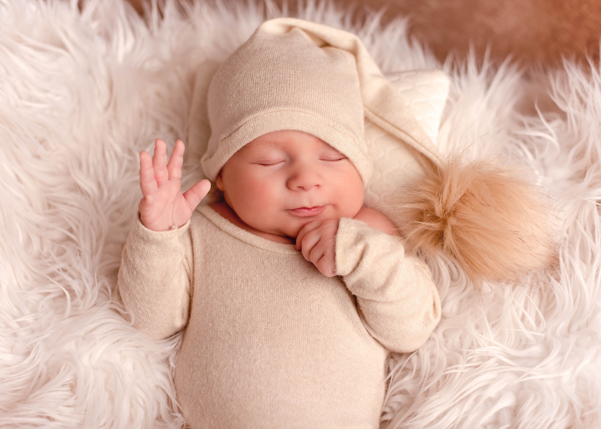 Śpiący noworodek w beżowym kombinezonie i czapce z pomponem, leżący na białym, puszystym futrze, z uniesioną dłonią.