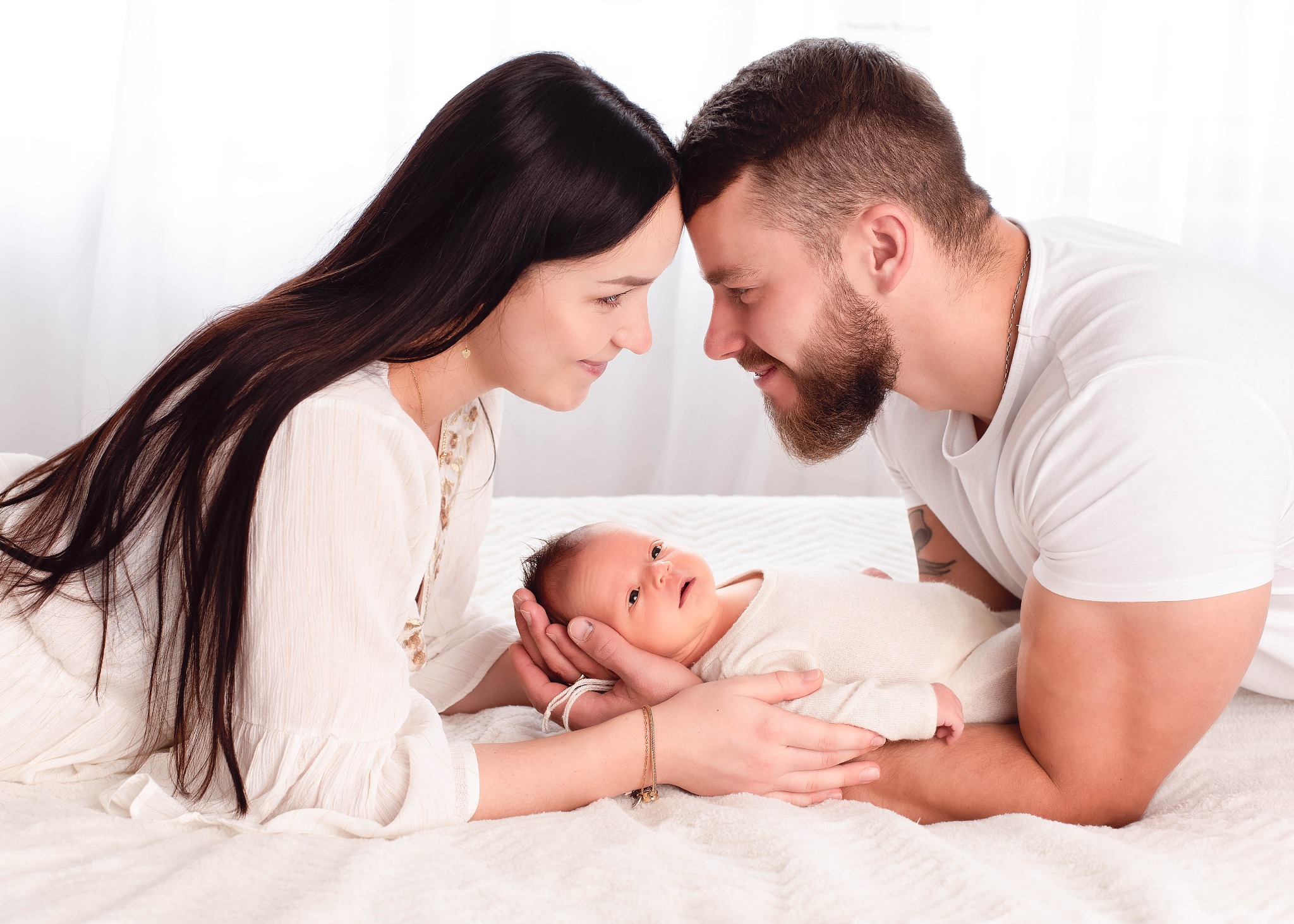 Rodzice patrzą na noworodka leżącego na białym kocu, czoła złączone, delikatne oświetlenie, naturalne kolory.