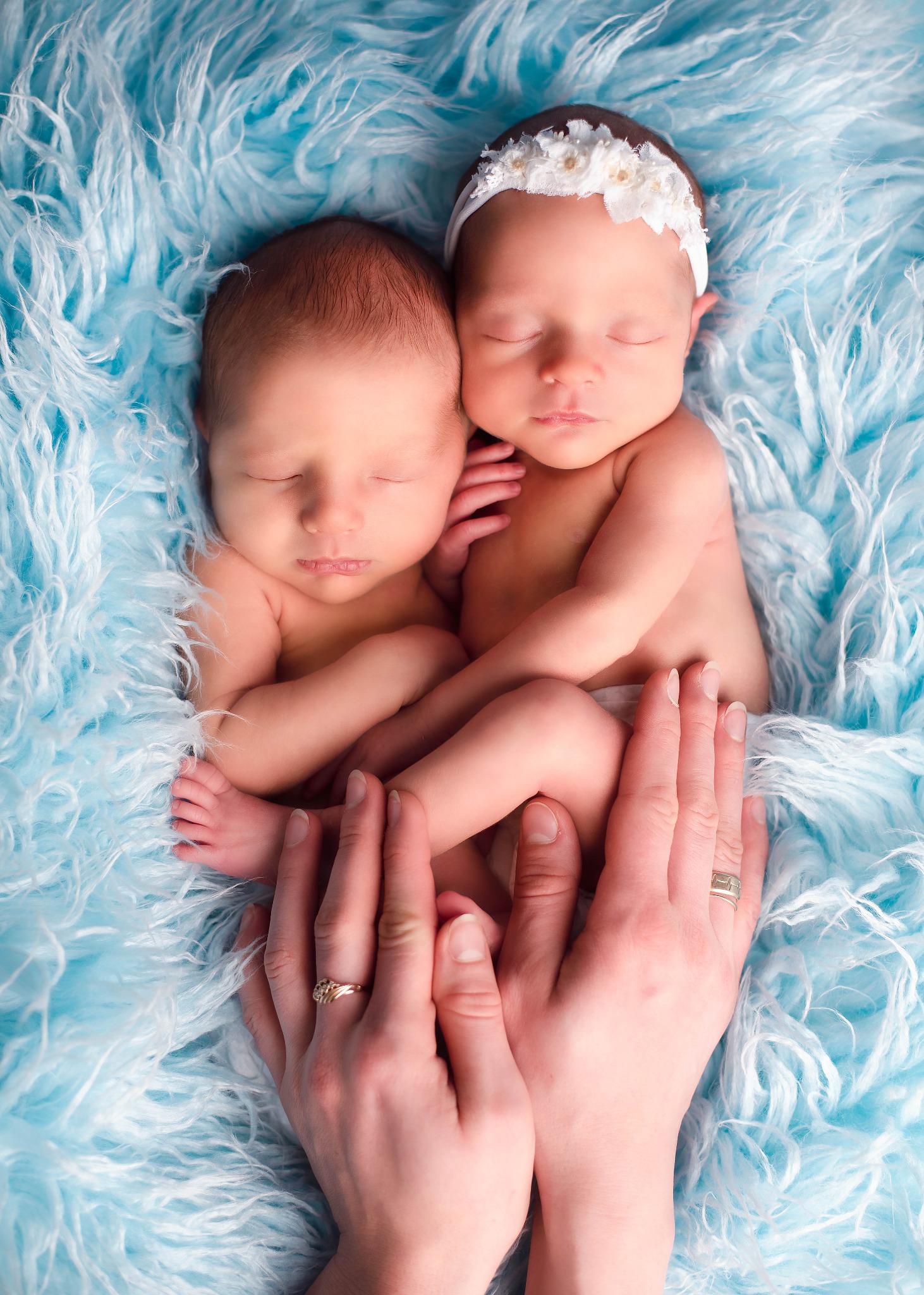 Dwoje śpiących noworodków, jedno z białą opaską na głowie, ułożone obok siebie na błękitnym, puszystym tle, dłonie rodziców delikatnie obejmujące dziecięce stópki, widoczne obrączki na palcach.