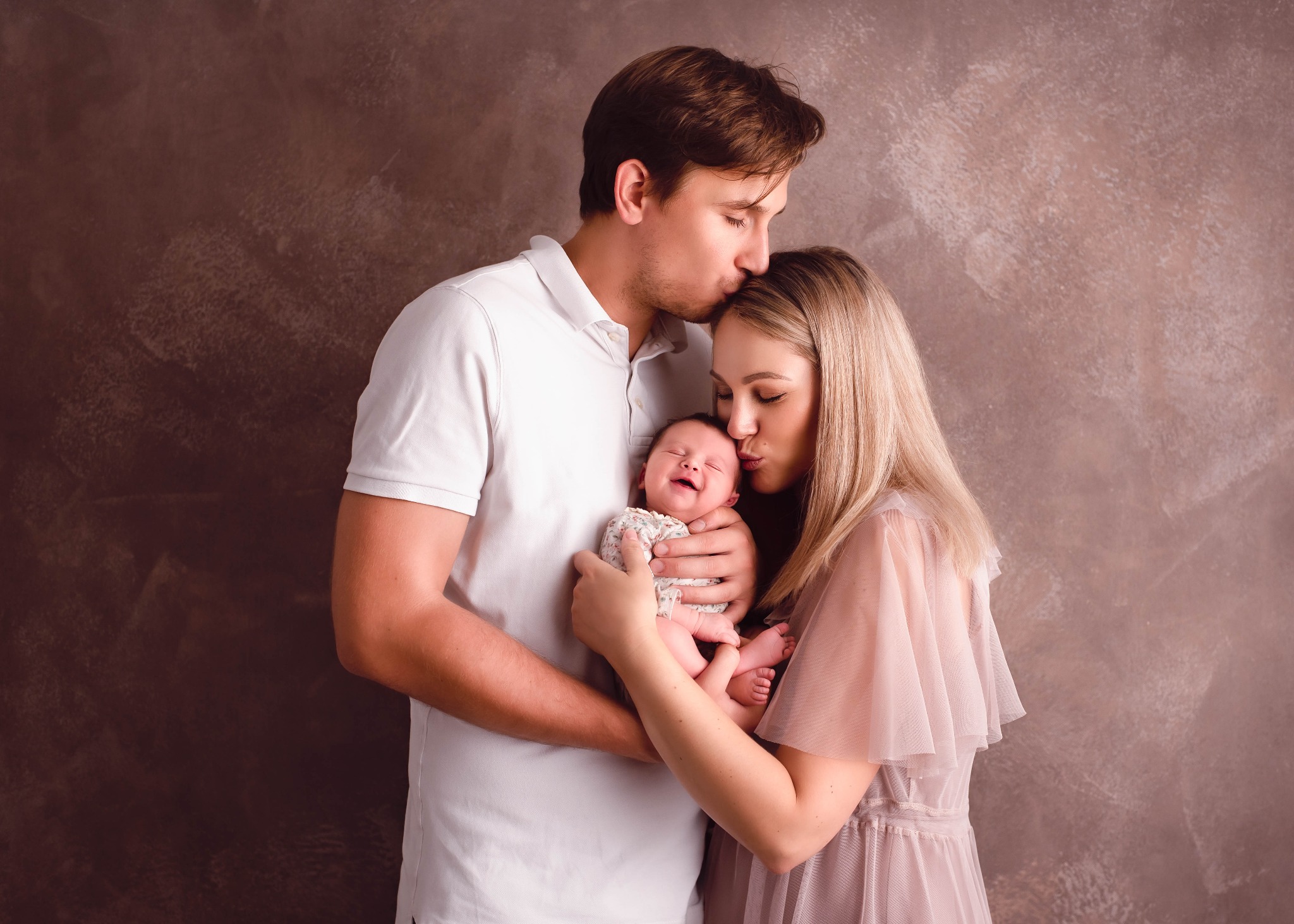 Rodzice całują nowo narodzone dziecko na tle teksturowanej ściany w odcieniach brązu. Dziecko uśmiecha się z zamkniętymi oczami.