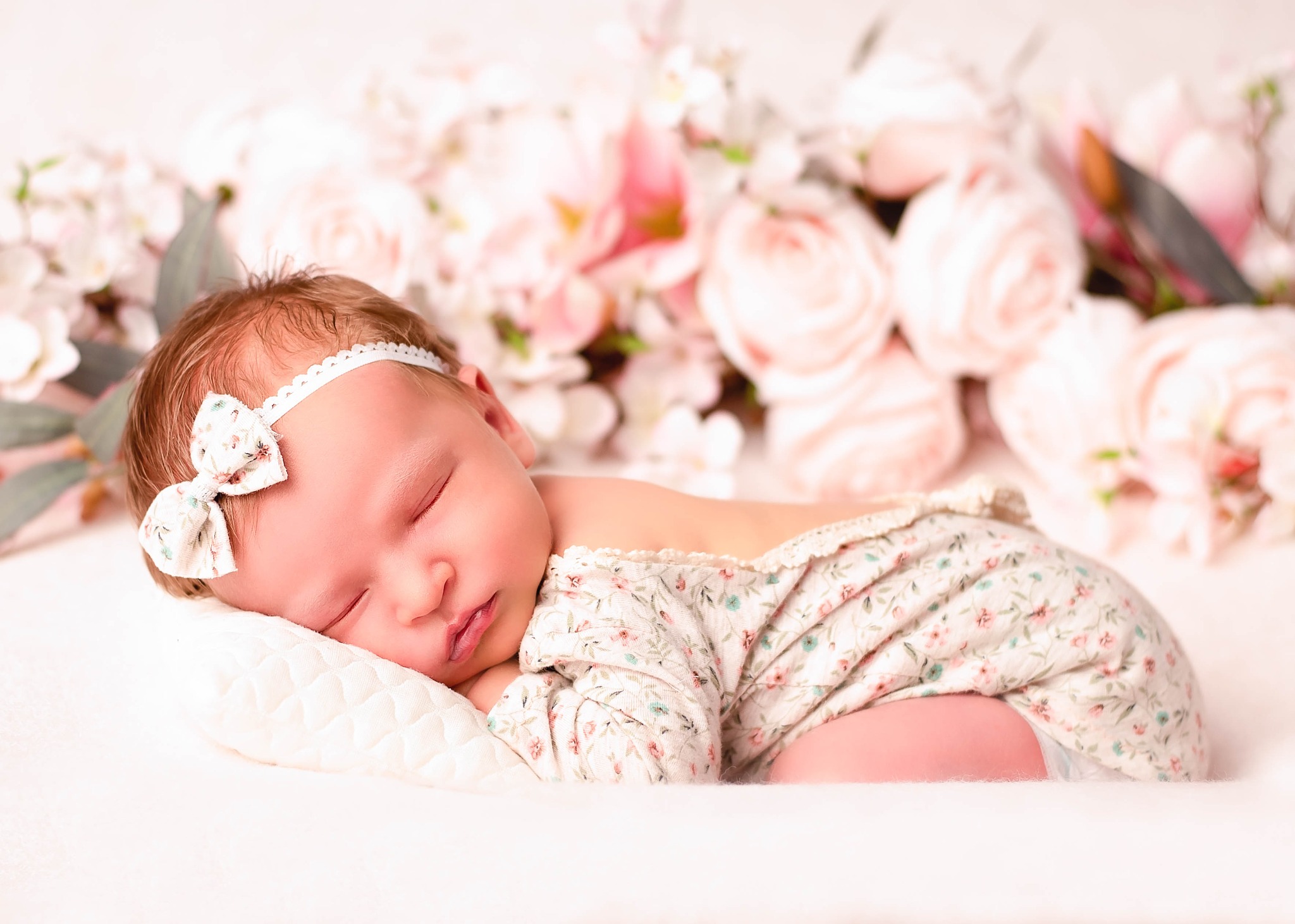 Śpiąca noworodka w kwiecistym ubranku i opasce z kokardką, ułożona na białej poduszce na tle rozmytych różowych kwiatów.