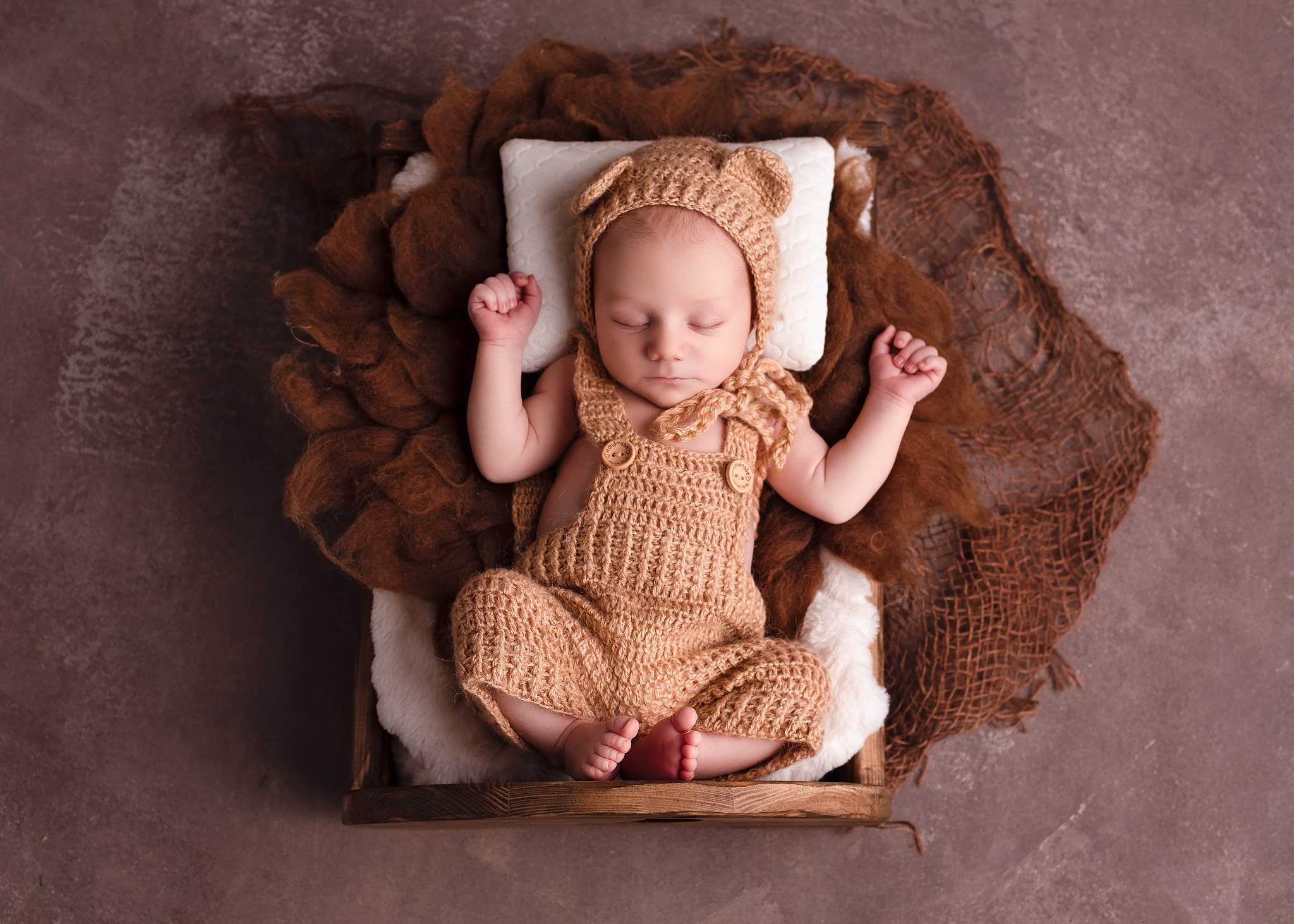 Noworodek śpiący na plecach w drewnianej skrzynce, ubrany w dziergany strój misia z uszami, na białej poduszce otoczonej brązową wełną.