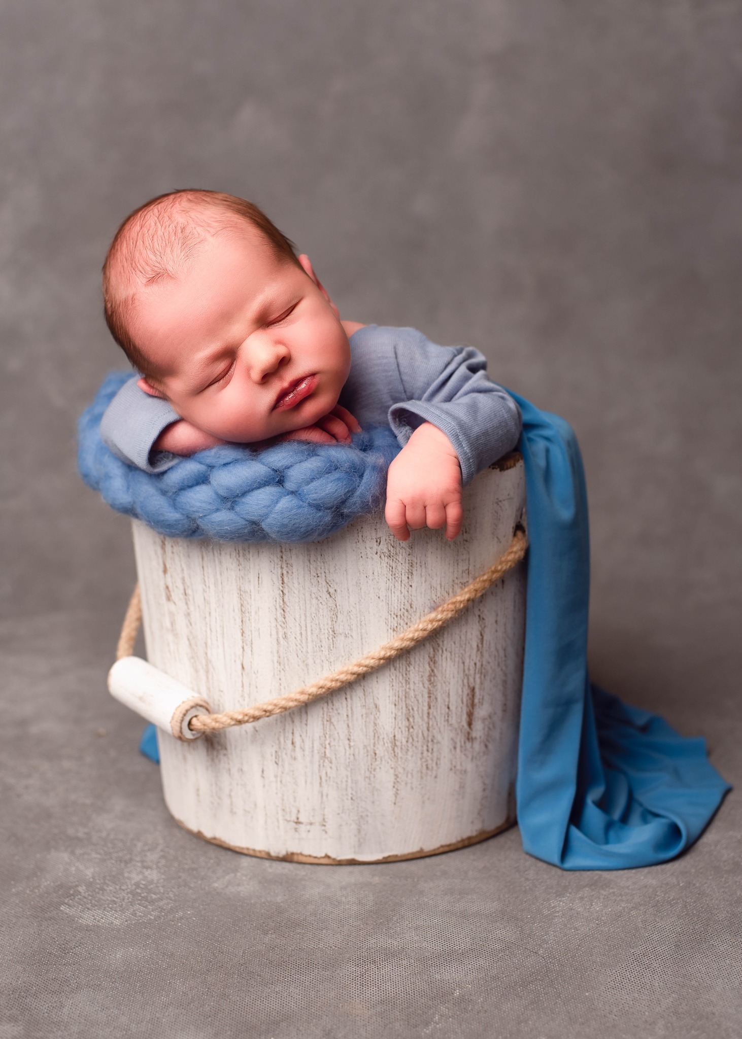Śpiący noworodek w błękitnym ubranku, ułożony w białym, stylizowanym wiaderku wypełnionym błękitną włóczką i otulony błękitnym materiałem, na szarym tle.