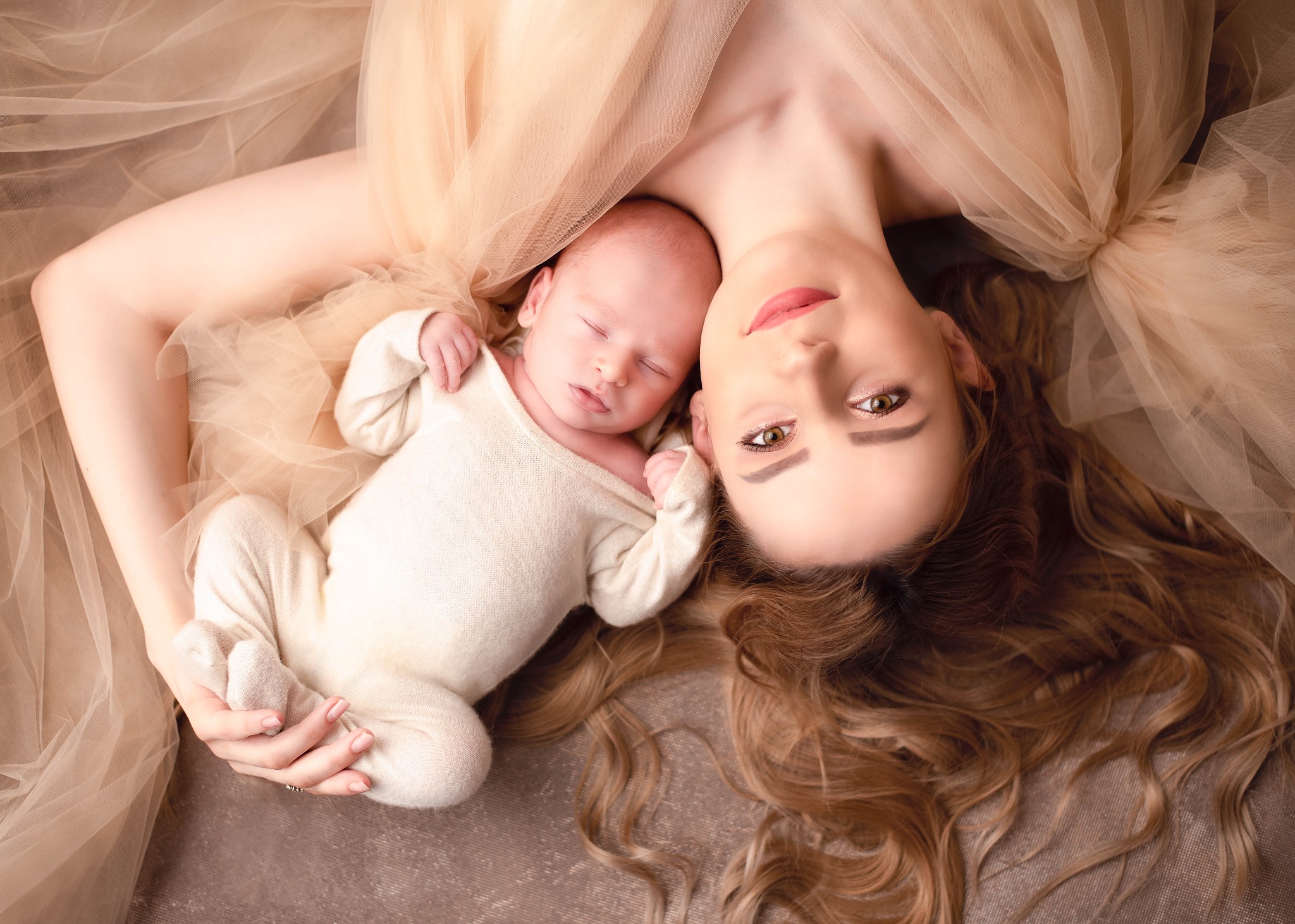 Matka leżąca na plecach z noworodkiem w beżowych ubrankach, otuleni tiulem w ciepłych odcieniach, delikatne oświetlenie podkreśla urodę.