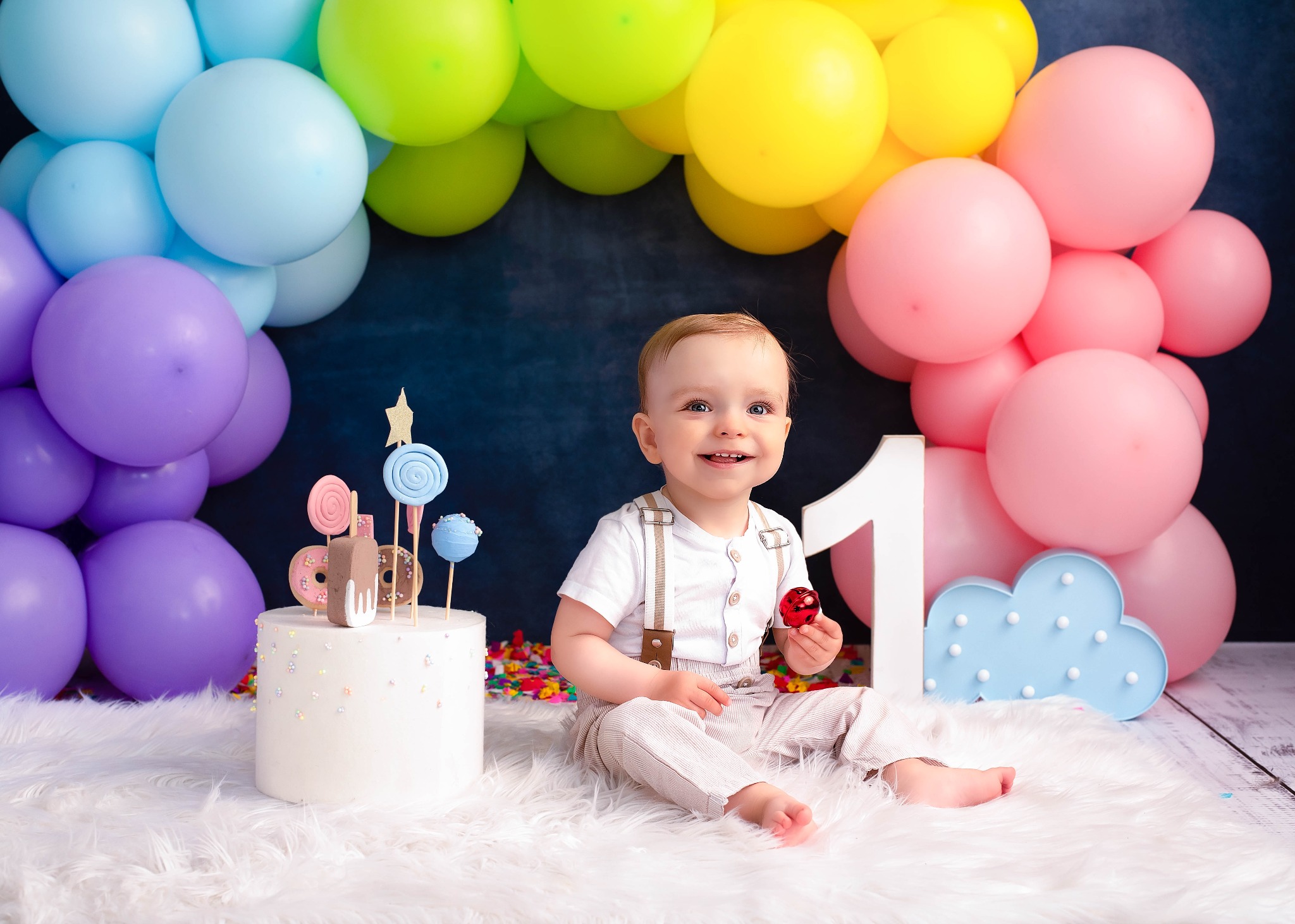 Uśmiechnięty roczny chłopiec w szelkach siedzi na białym futrze obok tortu z lizakami i cyfry '1', tło z kolorowych balonów.