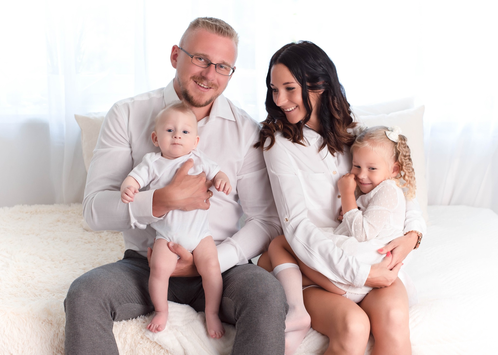 Uśmiechnięta rodzina: ojciec w okularach trzyma niemowlę w białym body, matka przytula starszą córkę z blond lokami i kokardami na białym, puszystym kocu.