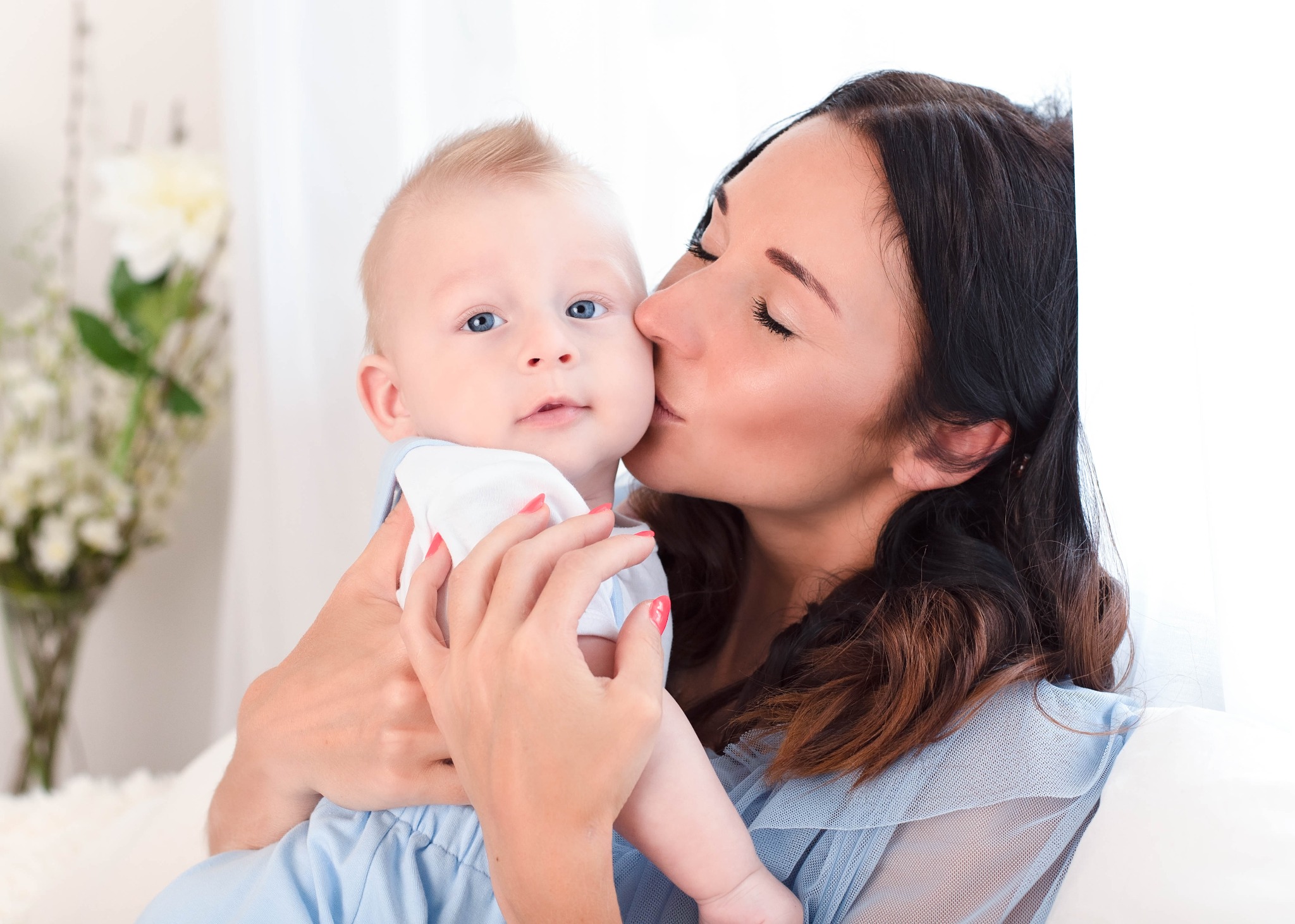 Matka całuje w policzek niemowlę o jasnych włosach i niebieskich oczach, ubrane w błękitny strój, na jasnym tle z kwiatami.