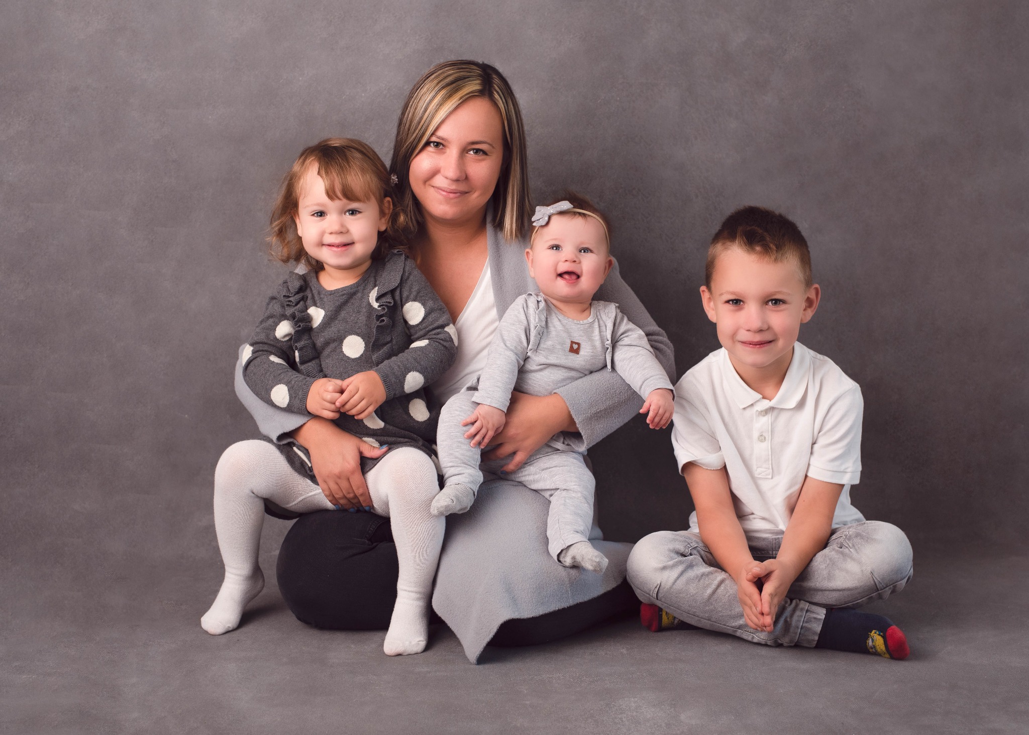 Uśmiechnięta mama z blond pasemkami pozuje z trójką dzieci na szarym tle w studio fotograficznym: dziewczynka w sukience w grochy na kolanach, niemowlę z kokardką na rękach i chłopiec w koszulce...
