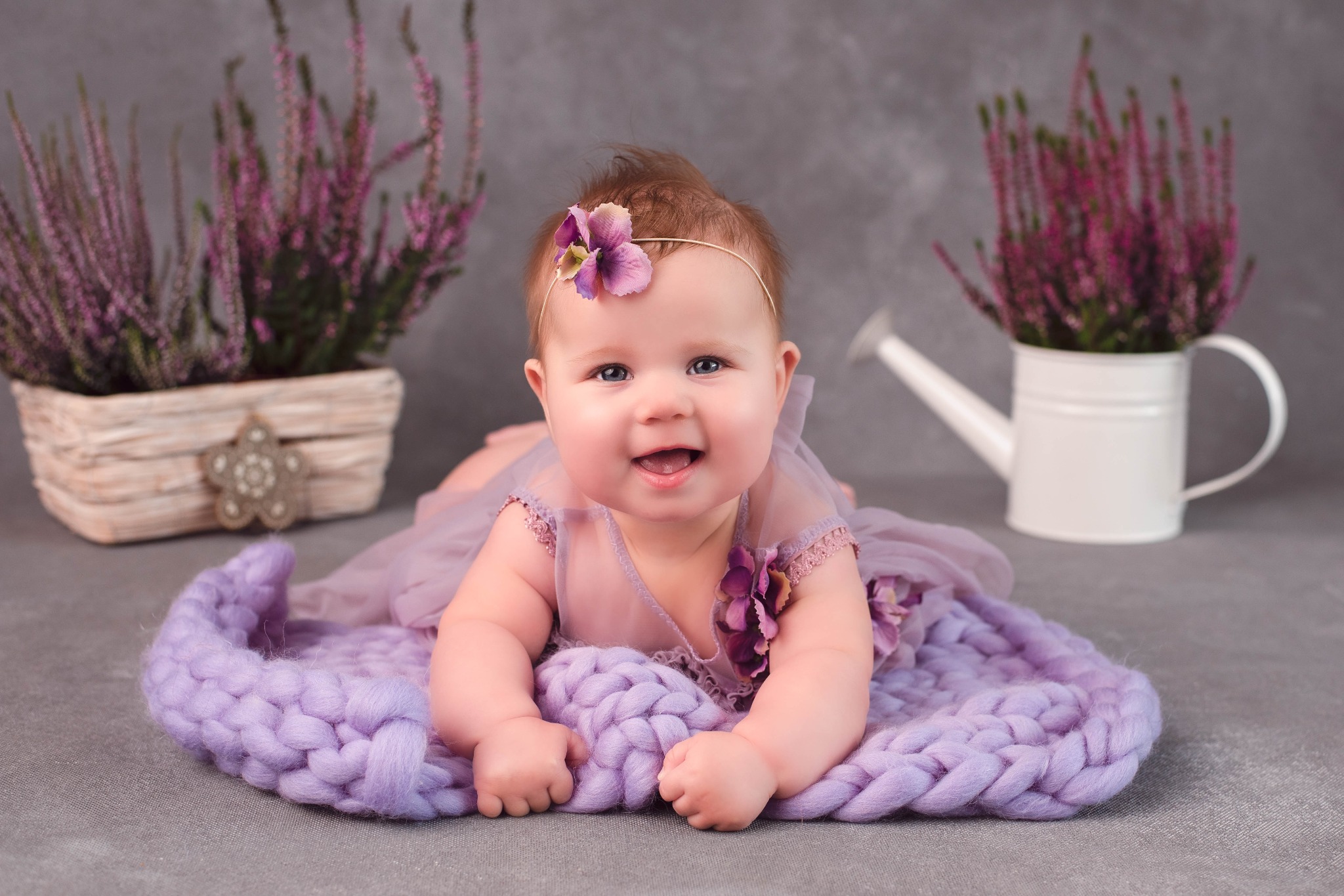 Uśmiechnięta niemowlęca dziewczynka leży na brzuchu na fioletowym, dzierganym kocyku, ubrana w tiulową sukienkę z kwiatami, z fioletowym kwiatkiem na głowie; w tle wrzosy w koszyku i konewka.