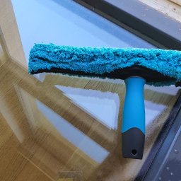 Window's Ninja - Mycie okna z użyciem niebieskiego czyścika z mikrofibry, pozostawiającego smugi na szybie, widoczne krople wody i ciemne obramowanie okna.