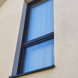 Window's Ninja - Nowoczesne okno z czarną ramą w elewacji budynku o jasnej, ziarnistej strukturze, widoczne refleksy nieba na szybie.