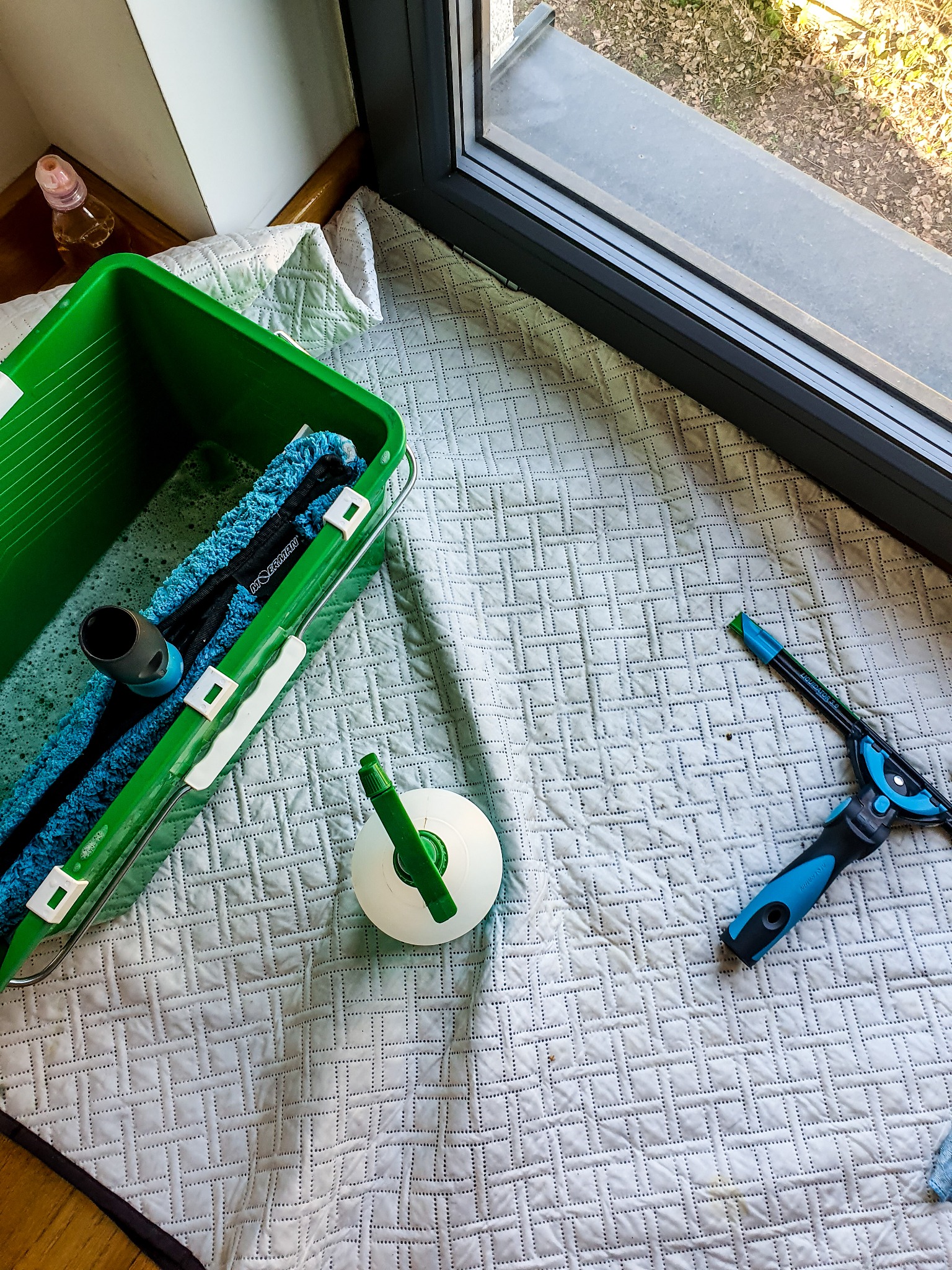 Przygotowanie do mycia okna: Wiadro z płynem, myjka z niebieską nakładką, spryskiwacz i ściągaczka do szyb leżą na białej kołdrze obok okna.