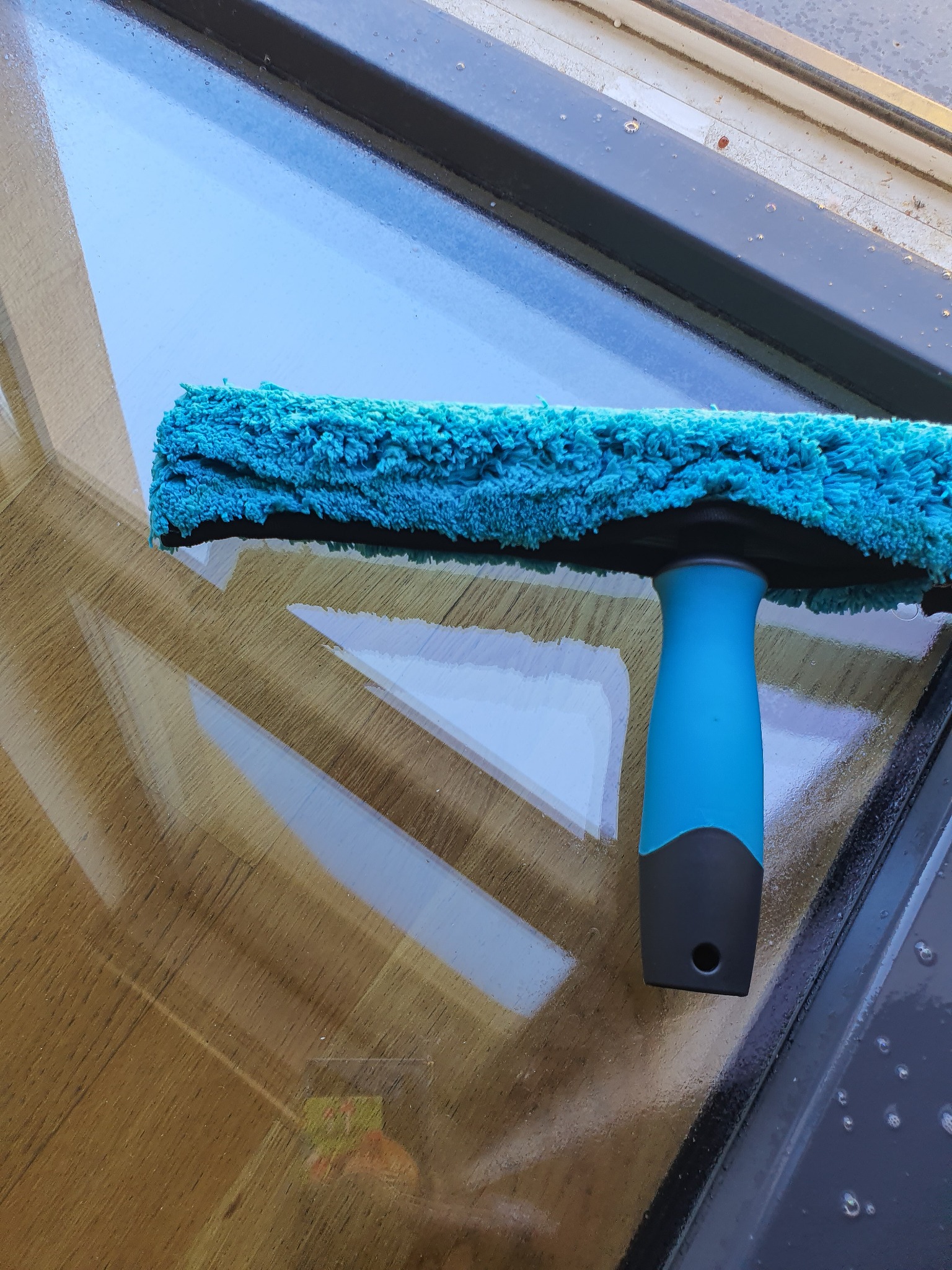 Mycie okna z użyciem niebieskiego czyścika z mikrofibry, pozostawiającego smugi na szybie, widoczne krople wody i ciemne obramowanie okna.