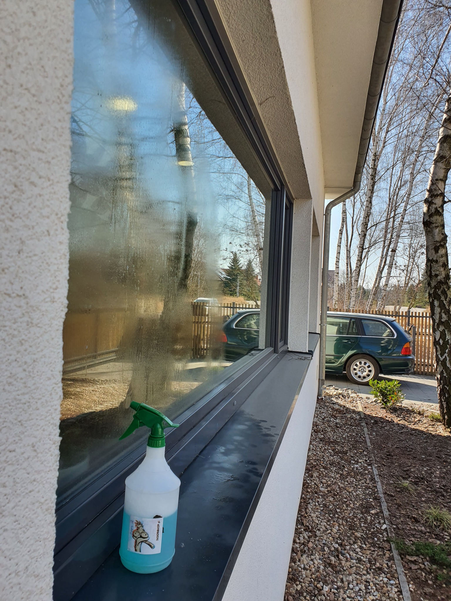 Okno z widocznymi smugami i zabrudzeniami przed myciem, obok stoi butelka ze spryskiwaczem i płynem do mycia szyb, w tle widoczny ogród i zaparkowany samochód kombi.