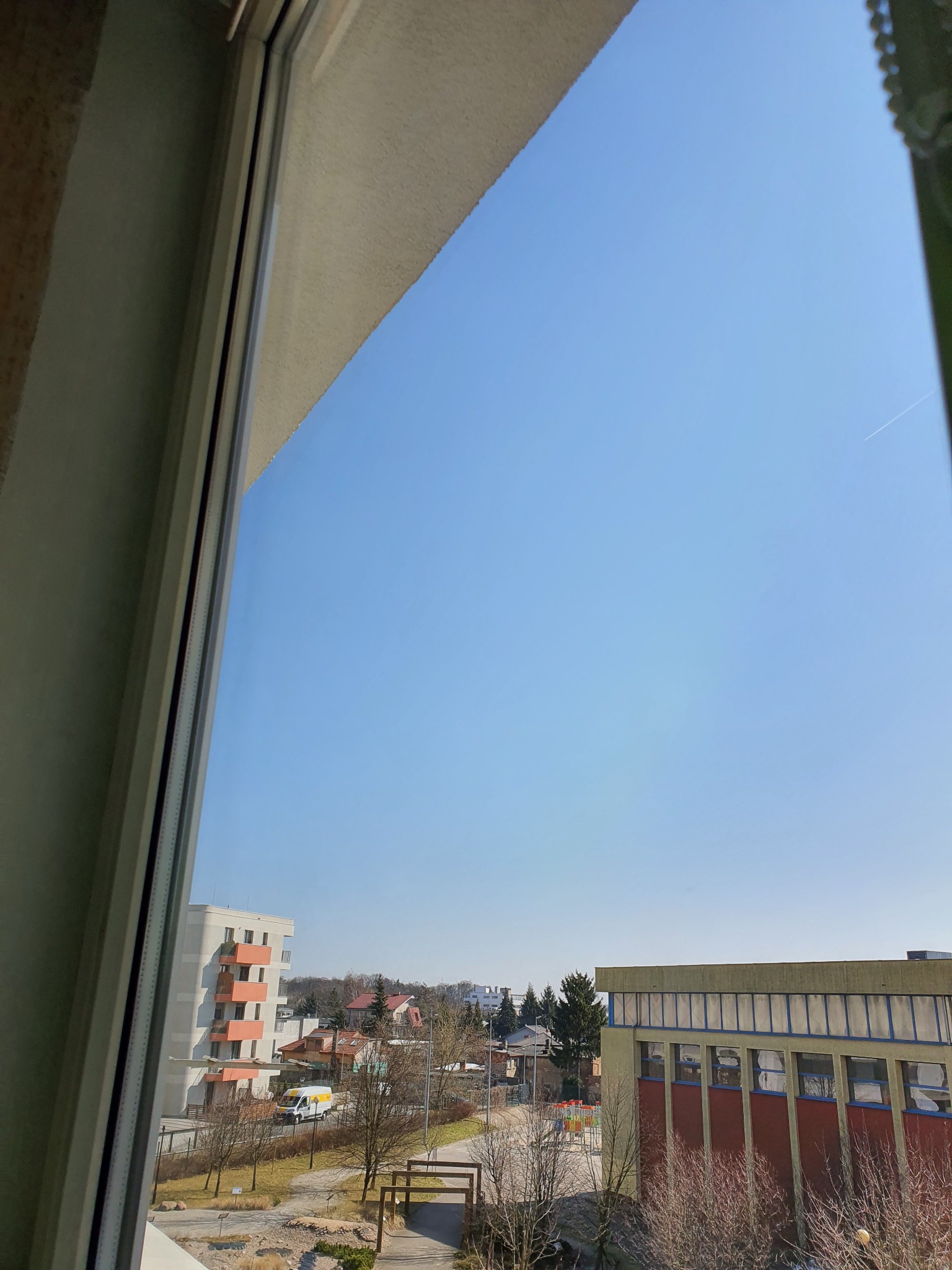 Widok z okna na błękitne niebo i budynki mieszkalne w słoneczny dzień, z widocznym śladem samolotu na niebie.