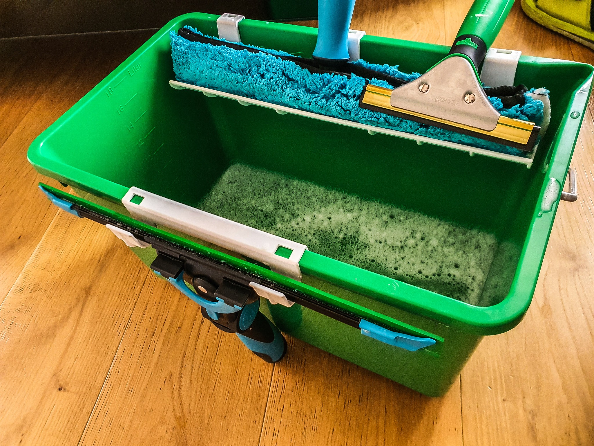 Zielone wiadro z wodą i pianą oraz różnymi narzędziami do mycia okien, w tym ściągaczki i myjka z niebieską mikrofibrą, leżące na drewnianej podłodze.