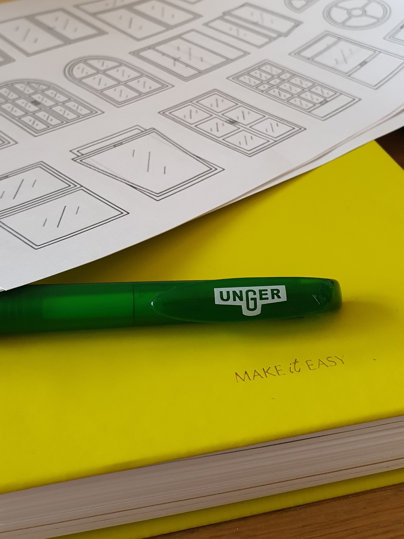 Szkicownik z rysunkami różnych typów okien, leżący na żółtym notatniku z zielonym pisakiem marki Unger.