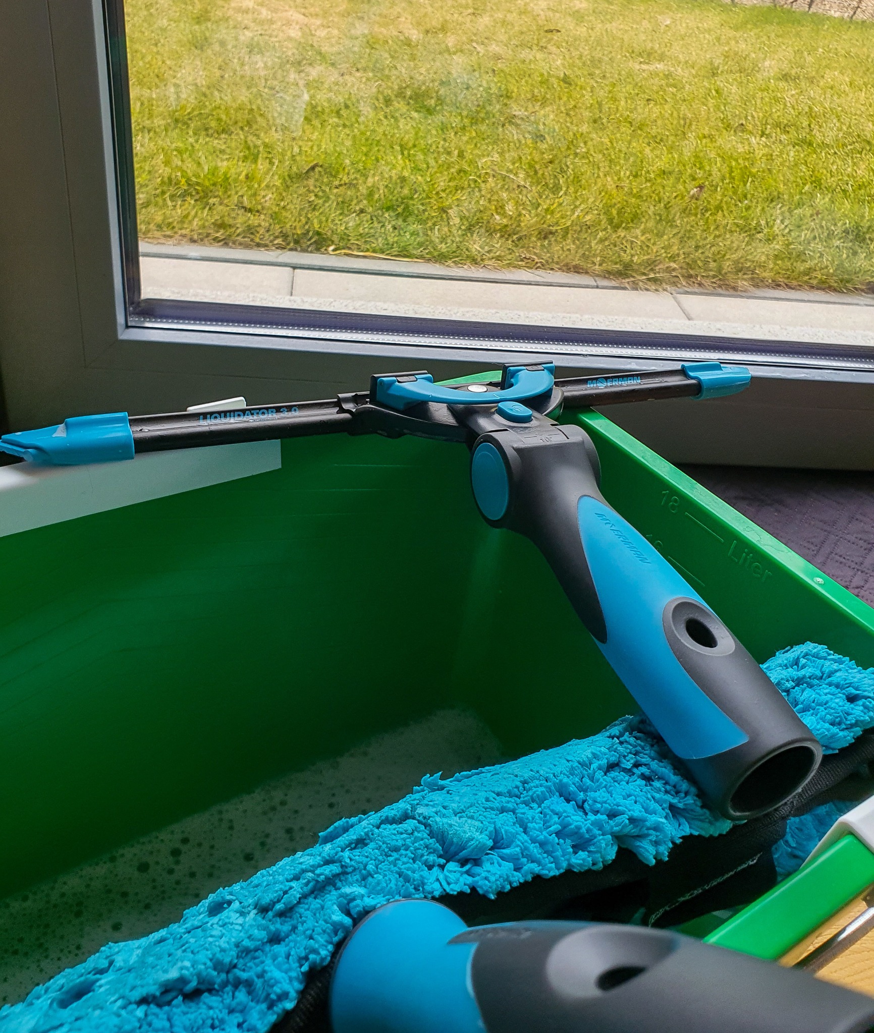 Profesjonalny zestaw do mycia okien: ściągaczka Liquidator 3.0 w zielonym wiadrze z wodą i płynem, widoczna niebieska ściereczka z mikrofibry oraz szyba okienna z widokiem na trawnik.
