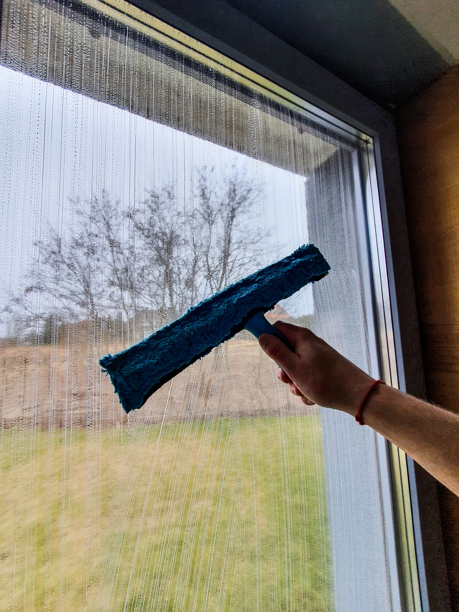 Mężczyzna czyści okno za pomocą niebieskiej myjki, widoczne smugi wody na szybie i drzewo w tle.