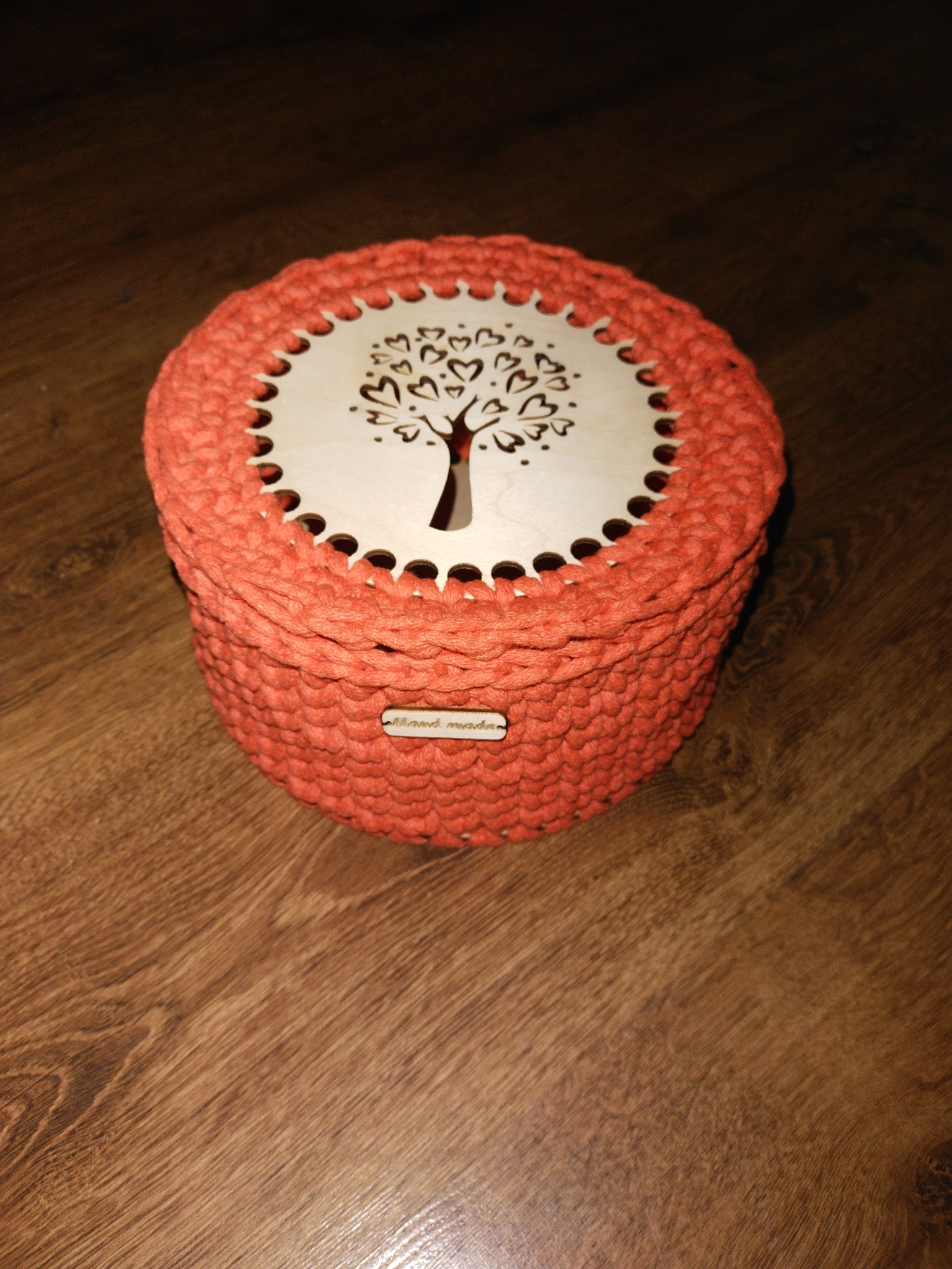 Pomarańczowy, dziergany koszyk z drewnianym wieczkiem z wyciętym motywem drzewa z sercami, leżący na drewnianej podłodze.