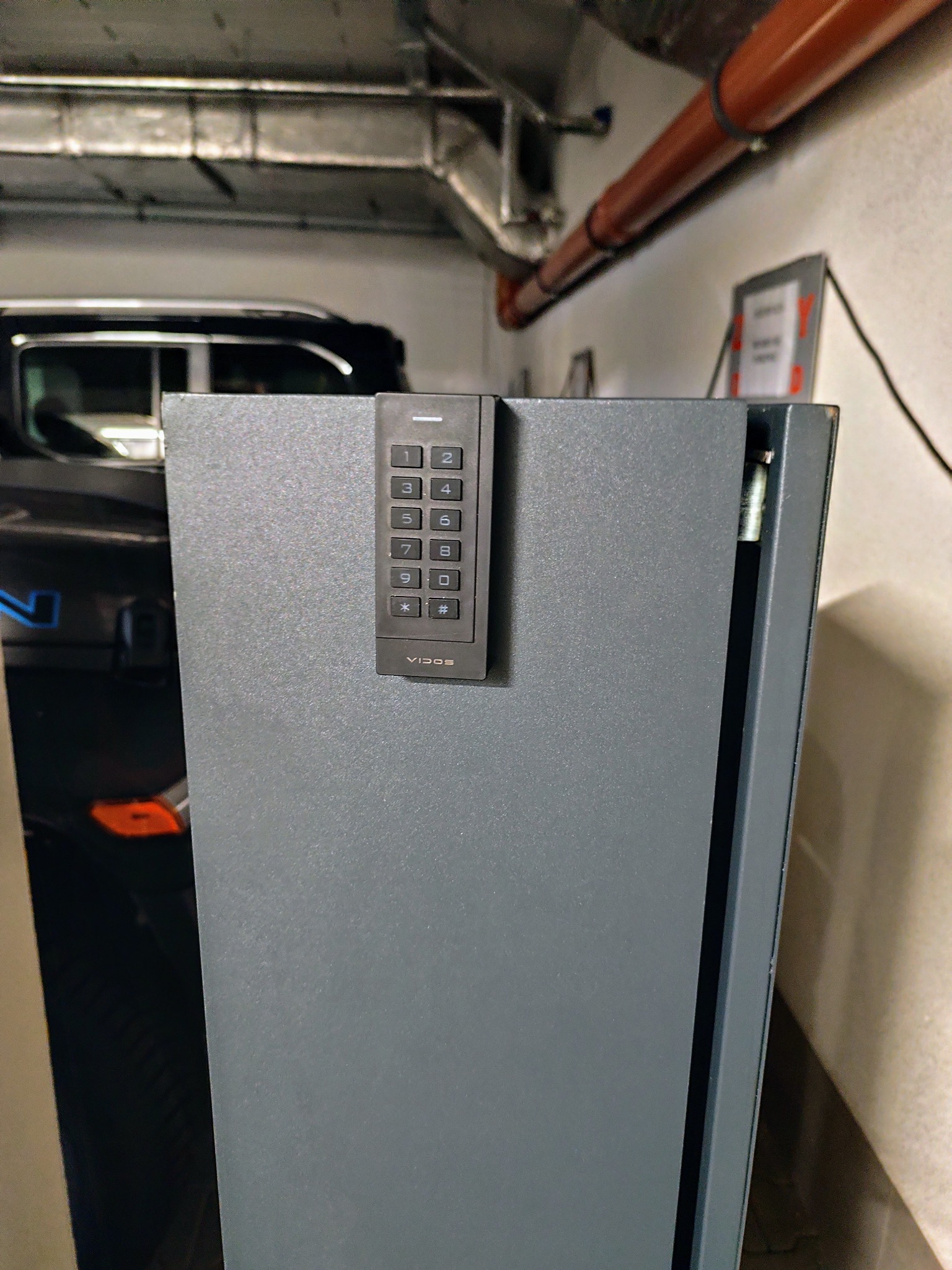 Szara obudowa ładowarki samochodowej z klawiaturą numeryczną Vidos w garażu podziemnym, z widocznym fragmentem samochodu w tle. Kontrola dostępu.