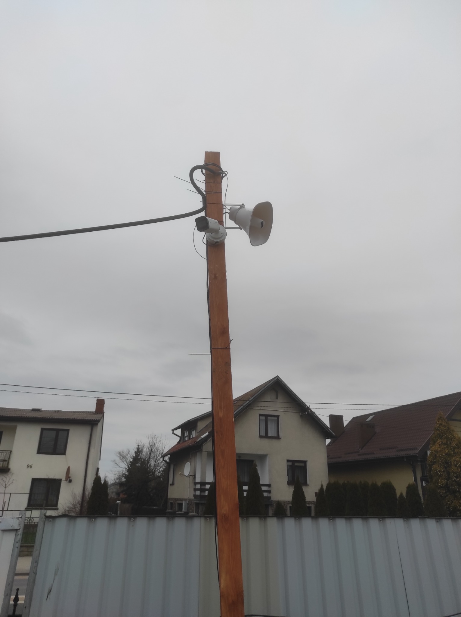 Słup z zamontowaną kamerą CCTV i głośnikiem tubowym, widoczne przewody, w tle budynki mieszkalne i pochmurne niebo.