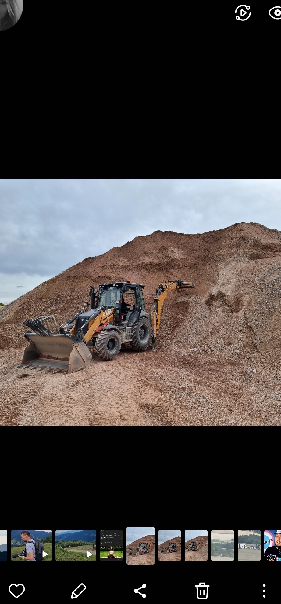 Żółto-czarna koparko-ładowarka Case na tle dużej pryzmy ziemi lub żwiru pod pochmurnym niebem. Widoczne ślady opon i nierówności terenu.