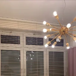 Jasne drewniane żaluzje poziome zamontowane w oknach, widoczne z wnętrza pomieszczenia, oświetlone przez nowoczesny, złoty żyrandol z żarówkami.